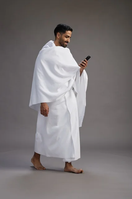 A portrait of an Arab Saudi Gulf man wearing Ihram clothing using a mobile phone, concept of Hajj and Umrah, full body, gray background.