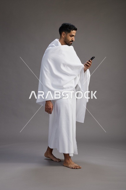 A portrait of an Arab Saudi Gulf man wearing Ihram clothing using a mobile phone, concept of Hajj and Umrah, full body, gray background.