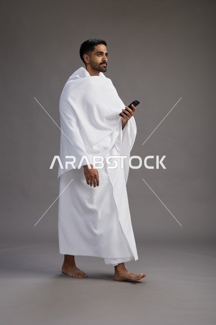 بورتريه لرجل عربي سعودي خليجي يرتدي ملابس الإحرام يستخدم الهاتف المحمول, مفهوم الحج والعمره , كامل الجسم, خلفيه رماديه