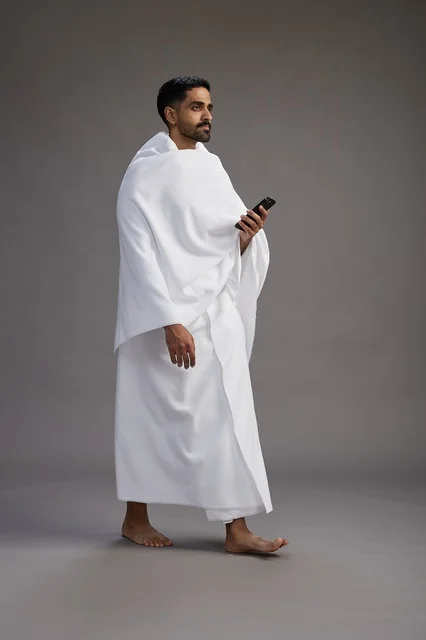 A portrait of an Arab Saudi Gulf man wearing Ihram clothing using a mobile phone, concept of Hajj and Umrah, full body, gray background.