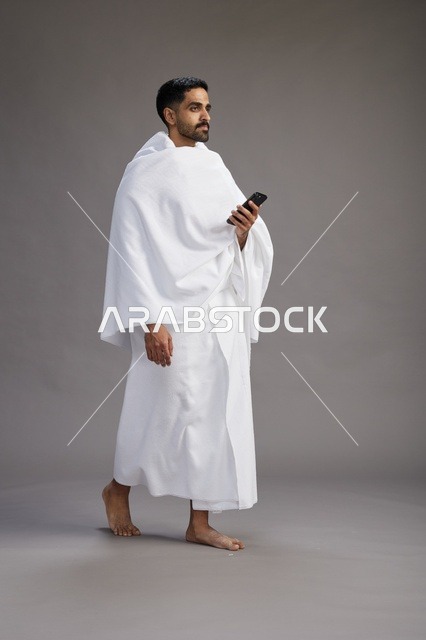 بورتريه لرجل عربي سعودي خليجي يرتدي ملابس الإحرام يستخدم الهاتف المحمول, مفهوم الحج والعمره , كامل الجسم, خلفيه رماديه