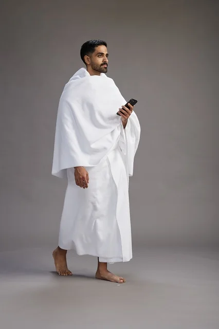 بورتريه لرجل عربي سعودي خليجي يرتدي ملابس الإحرام يستخدم الهاتف المحمول, مفهوم الحج والعمره , كامل الجسم, خلفيه رماديه