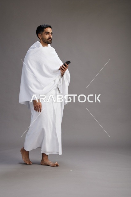 بورتريه لرجل عربي سعودي خليجي يرتدي ملابس الإحرام يستخدم الهاتف المحمول, مفهوم الحج والعمره , كامل الجسم, خلفيه رماديه