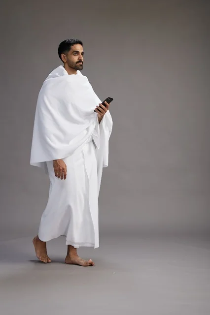 A portrait of an Arab Saudi Gulf man wearing Ihram clothing using a mobile phone, concept of Hajj and Umrah, full body, gray background.