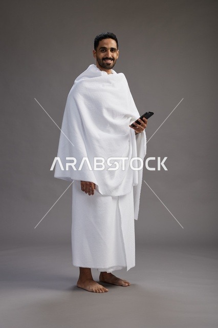 بورتريه لرجل عربي سعودي خليجي يرتدي ملابس الإحرام يستخدم الهاتف المحمول, مفهوم الحج والعمره , كامل الجسم, خلفيه رماديه