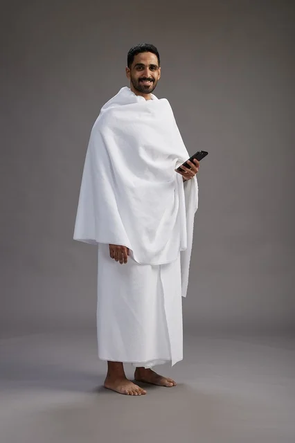 A portrait of an Arab Saudi Gulf man wearing Ihram clothing using a mobile phone, concept of Hajj and Umrah, full body, gray background.