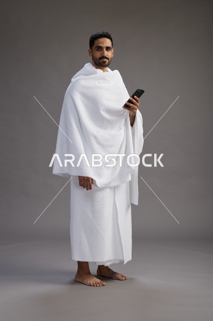 بورتريه لرجل عربي سعودي خليجي يرتدي ملابس الإحرام يستخدم الهاتف المحمول, مفهوم الحج والعمره , كامل الجسم, خلفيه رماديه