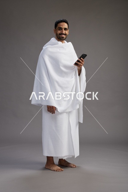 بورتريه لرجل عربي سعودي خليجي يرتدي ملابس الإحرام يستخدم الهاتف المحمول, مفهوم الحج والعمره , كامل الجسم, خلفيه رماديه