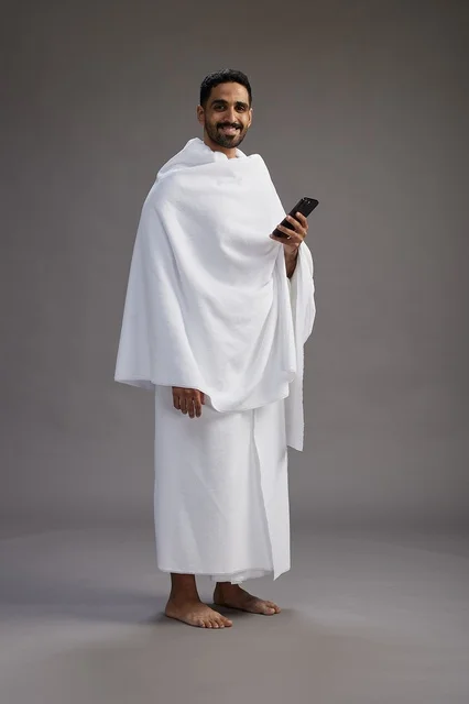 A portrait of an Arab Saudi Gulf man wearing Ihram clothing using a mobile phone, concept of Hajj and Umrah, full body, gray background.