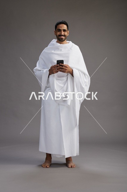 بورتريه لرجل عربي سعودي خليجي يرتدي ملابس الإحرام يستخدم الهاتف المحمول, مفهوم الحج والعمره , كامل الجسم, خلفيه رماديه