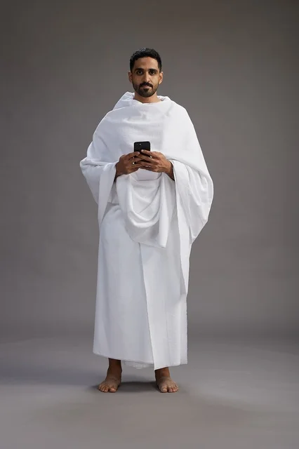 A portrait of an Arab Saudi Gulf man wearing Ihram clothing using a mobile phone, concept of Hajj and Umrah, full body, gray background.
