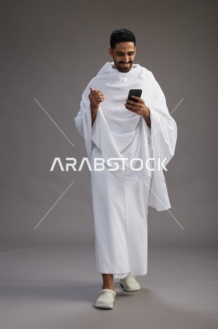 A portrait of an Arab Saudi Gulf man wearing Ihram clothing using a mobile phone, concept of Hajj and Umrah, full body, gray background.