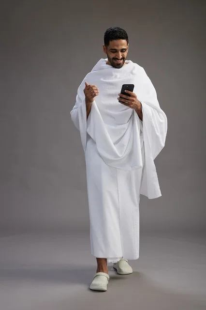 A portrait of an Arab Saudi Gulf man wearing Ihram clothing using a mobile phone, concept of Hajj and Umrah, full body, gray background.