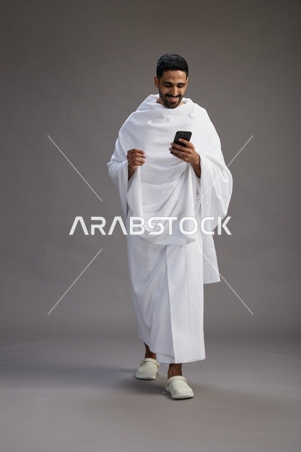 A portrait of an Arab Saudi Gulf man wearing Ihram clothing using a mobile phone, concept of Hajj and Umrah, full body, gray background.