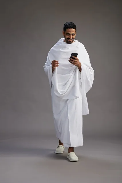 A portrait of an Arab Saudi Gulf man wearing Ihram clothing using a mobile phone, concept of Hajj and Umrah, full body, gray background.