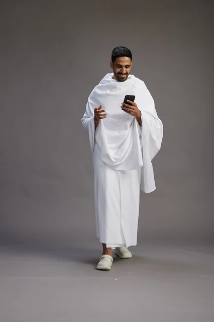 A portrait of an Arab Saudi Gulf man wearing Ihram clothing using a mobile phone, concept of Hajj and Umrah, full body, gray background.