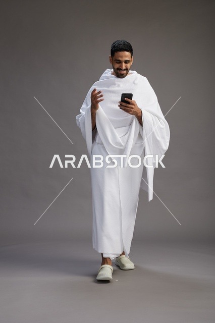 بورتريه لرجل عربي سعودي خليجي يرتدي ملابس الإحرام يستخدم الهاتف المحمول, مفهوم الحج والعمره , كامل الجسم, خلفيه رماديه