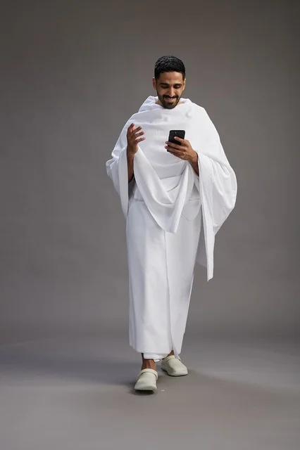 بورتريه لرجل عربي سعودي خليجي يرتدي ملابس الإحرام يستخدم الهاتف المحمول, مفهوم الحج والعمره , كامل الجسم, خلفيه رماديه