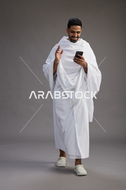 بورتريه لرجل عربي سعودي خليجي يرتدي ملابس الإحرام يستخدم الهاتف المحمول, مفهوم الحج والعمره , كامل الجسم, خلفيه رماديه