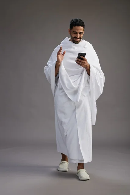 A portrait of an Arab Saudi Gulf man wearing Ihram clothing using a mobile phone, concept of Hajj and Umrah, full body, gray background.