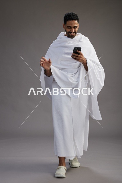 A portrait of an Arab Saudi Gulf man wearing Ihram clothing using a mobile phone, concept of Hajj and Umrah, full body, gray background.
