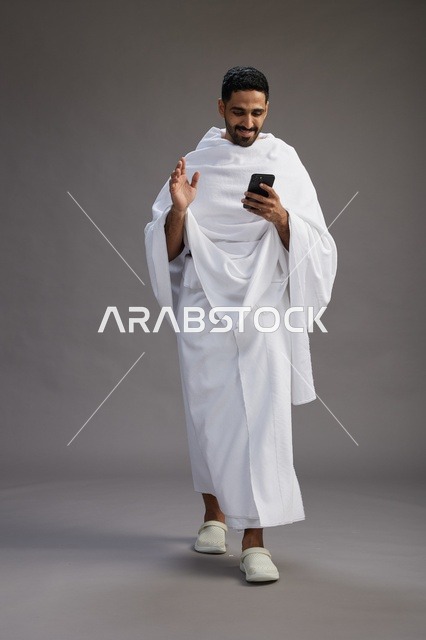 بورتريه لرجل عربي سعودي خليجي يرتدي ملابس الإحرام يستخدم الهاتف المحمول, مفهوم الحج والعمره , كامل الجسم, خلفيه رماديه