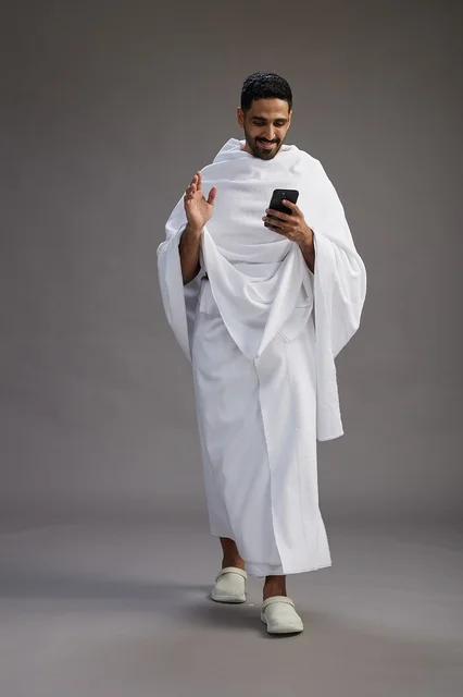 A portrait of an Arab Saudi Gulf man wearing Ihram clothing using a mobile phone, concept of Hajj and Umrah, full body, gray background.