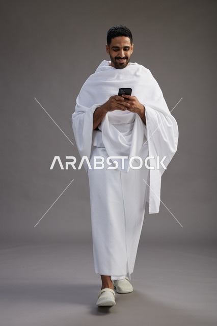 بورتريه لرجل عربي سعودي خليجي يرتدي ملابس الإحرام يستخدم الهاتف المحمول, مفهوم الحج والعمره , كامل الجسم, خلفيه رماديه