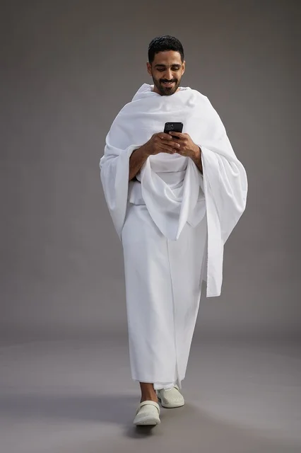 بورتريه لرجل عربي سعودي خليجي يرتدي ملابس الإحرام يستخدم الهاتف المحمول, مفهوم الحج والعمره , كامل الجسم, خلفيه رماديه