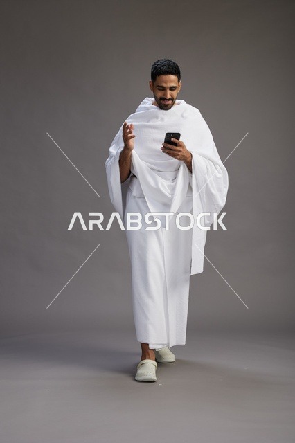 بورتريه لرجل عربي سعودي خليجي يرتدي ملابس الإحرام يستخدم الهاتف المحمول, مفهوم الحج والعمره , كامل الجسم, خلفيه رماديه
