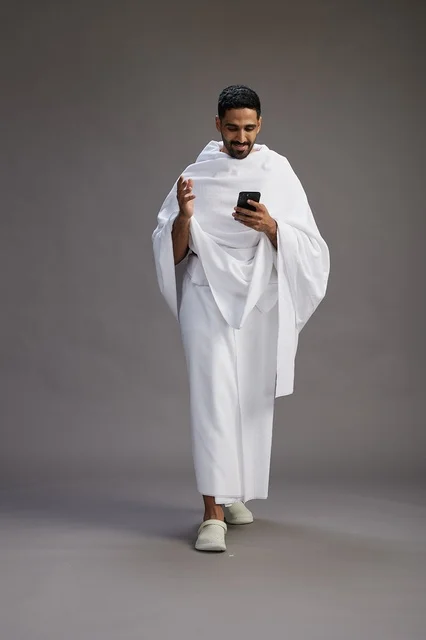 بورتريه لرجل عربي سعودي خليجي يرتدي ملابس الإحرام يستخدم الهاتف المحمول, مفهوم الحج والعمره , كامل الجسم, خلفيه رماديه