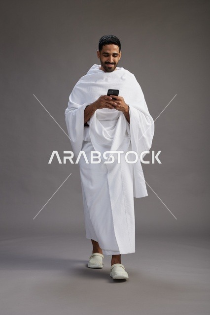 بورتريه لرجل عربي سعودي خليجي يرتدي ملابس الإحرام يستخدم الهاتف المحمول, مفهوم الحج والعمره , كامل الجسم, خلفيه رماديه