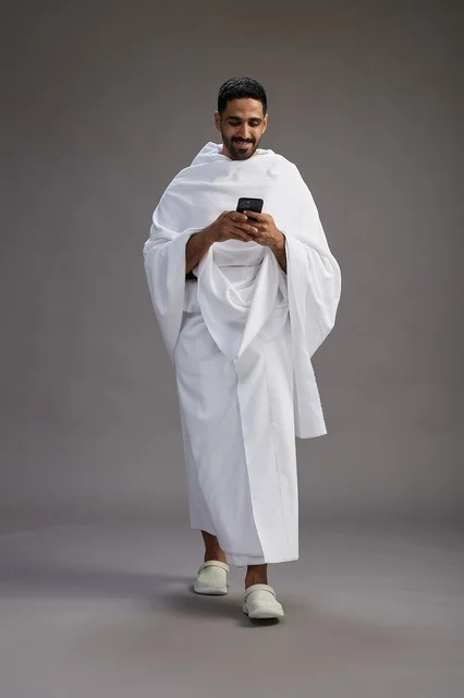بورتريه لرجل عربي سعودي خليجي يرتدي ملابس الإحرام يستخدم الهاتف المحمول, مفهوم الحج والعمره , كامل الجسم, خلفيه رماديه