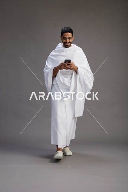 بورتريه لرجل عربي سعودي خليجي يرتدي ملابس الإحرام يستخدم الهاتف المحمول, مفهوم الحج والعمره , كامل الجسم, خلفيه رماديه
