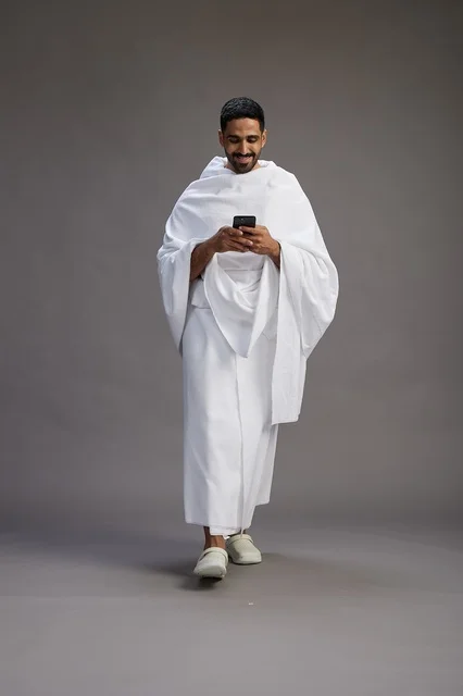 A portrait of an Arab Saudi Gulf man wearing Ihram clothing using a mobile phone, concept of Hajj and Umrah, full body, gray background.