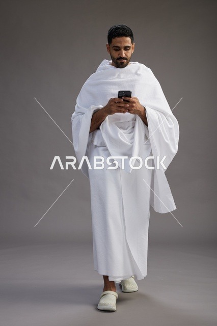 بورتريه لرجل عربي سعودي خليجي يرتدي ملابس الإحرام يستخدم الهاتف المحمول, مفهوم الحج والعمره , كامل الجسم, خلفيه رماديه