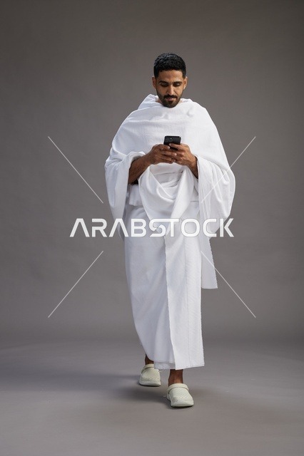 بورتريه لرجل عربي سعودي خليجي يرتدي ملابس الإحرام يستخدم الهاتف المحمول, مفهوم الحج والعمره , كامل الجسم, خلفيه رماديه