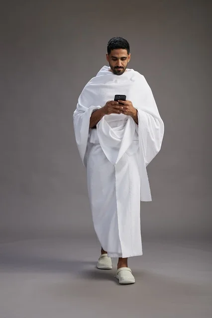 A portrait of an Arab Saudi Gulf man wearing Ihram clothing using a mobile phone, concept of Hajj and Umrah, full body, gray background.