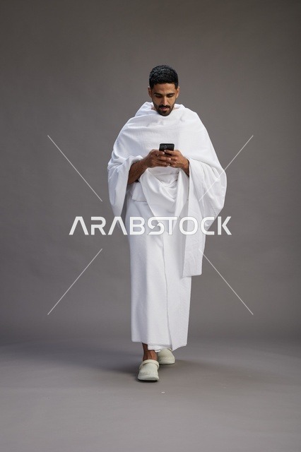 بورتريه لرجل عربي سعودي خليجي يرتدي ملابس الإحرام يستخدم الهاتف المحمول, مفهوم الحج والعمره , كامل الجسم, خلفيه رماديه