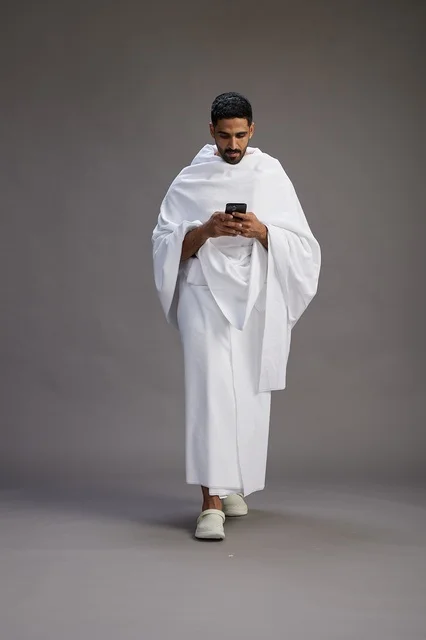 A portrait of an Arab Saudi Gulf man wearing Ihram clothing using a mobile phone, concept of Hajj and Umrah, full body, gray background.