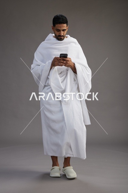 بورتريه لرجل عربي سعودي خليجي يرتدي ملابس الإحرام يستخدم الهاتف المحمول, مفهوم الحج والعمره , كامل الجسم, خلفيه رماديه