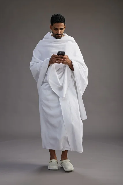 A portrait of an Arab Saudi Gulf man wearing Ihram clothing using a mobile phone, concept of Hajj and Umrah, full body, gray background.