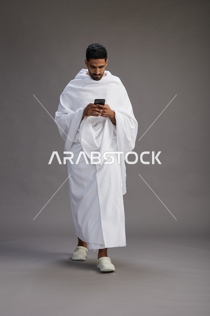 بورتريه لرجل عربي سعودي خليجي يرتدي ملابس الإحرام يستخدم الهاتف المحمول, مفهوم الحج والعمره , كامل الجسم, خلفيه رماديه