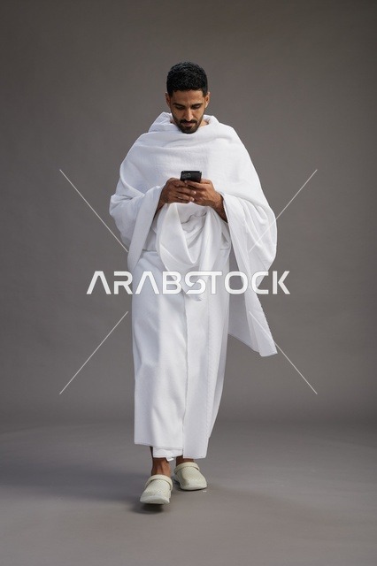 بورتريه لرجل عربي سعودي خليجي يرتدي ملابس الإحرام يستخدم الهاتف المحمول, مفهوم الحج والعمره , كامل الجسم, خلفيه رماديه