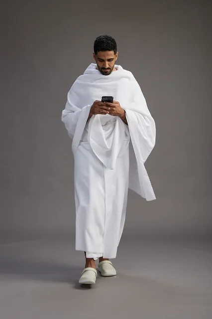 A portrait of an Arab Saudi Gulf man wearing Ihram clothing using a mobile phone, concept of Hajj and Umrah, full body, gray background.