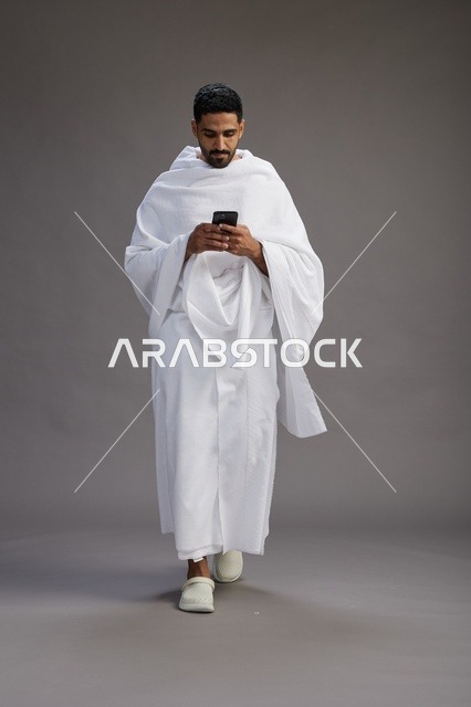 بورتريه لرجل عربي سعودي خليجي يرتدي ملابس الإحرام يستخدم الهاتف المحمول, مفهوم الحج والعمره , كامل الجسم, خلفيه رماديه