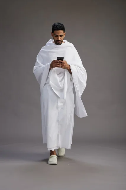 A portrait of an Arab Saudi Gulf man wearing Ihram clothing using a mobile phone, concept of Hajj and Umrah, full body, gray background. A portrait of an Arab Saudi Gulf man wearing Ihram clothing using a mobile phone, concept of Hajj and Umrah, full body, gray background.