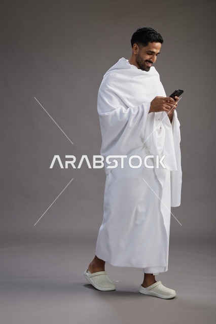 بورتريه لرجل عربي سعودي خليجي يرتدي ملابس الإحرام يستخدم الهاتف المحمول, مفهوم الحج والعمره , كامل الجسم, خلفيه رماديه