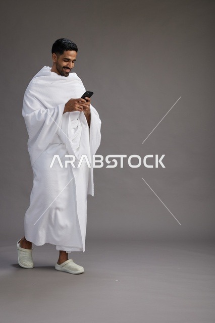 A portrait of an Arab Saudi Gulf man wearing Ihram clothing using a mobile phone, concept of Hajj and Umrah, full body, gray background.