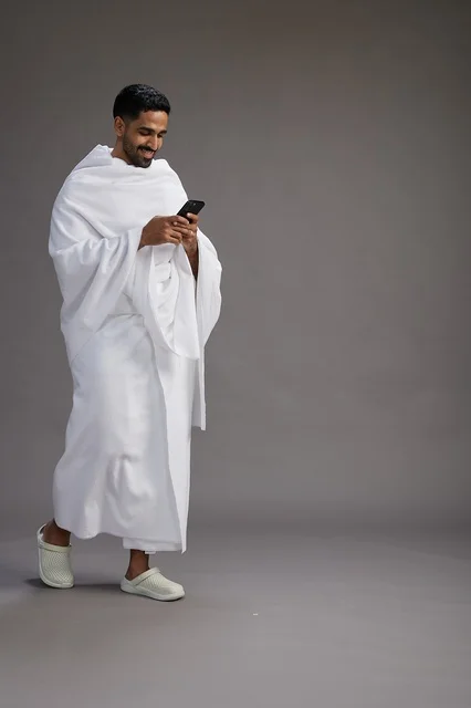 A portrait of an Arab Saudi Gulf man wearing Ihram clothing using a mobile phone, concept of Hajj and Umrah, full body, gray background.